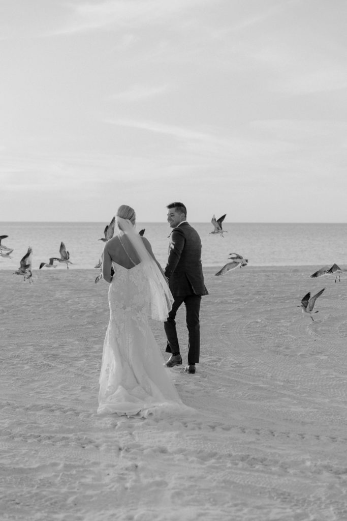 Wedding Photographer, bride and groom walk hand in hand at the beach through the sand