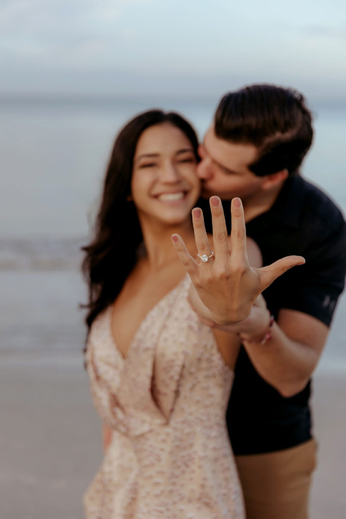 Elopement Photographer, man kisses woman on the cheek at the beach, she shows off engagement ring